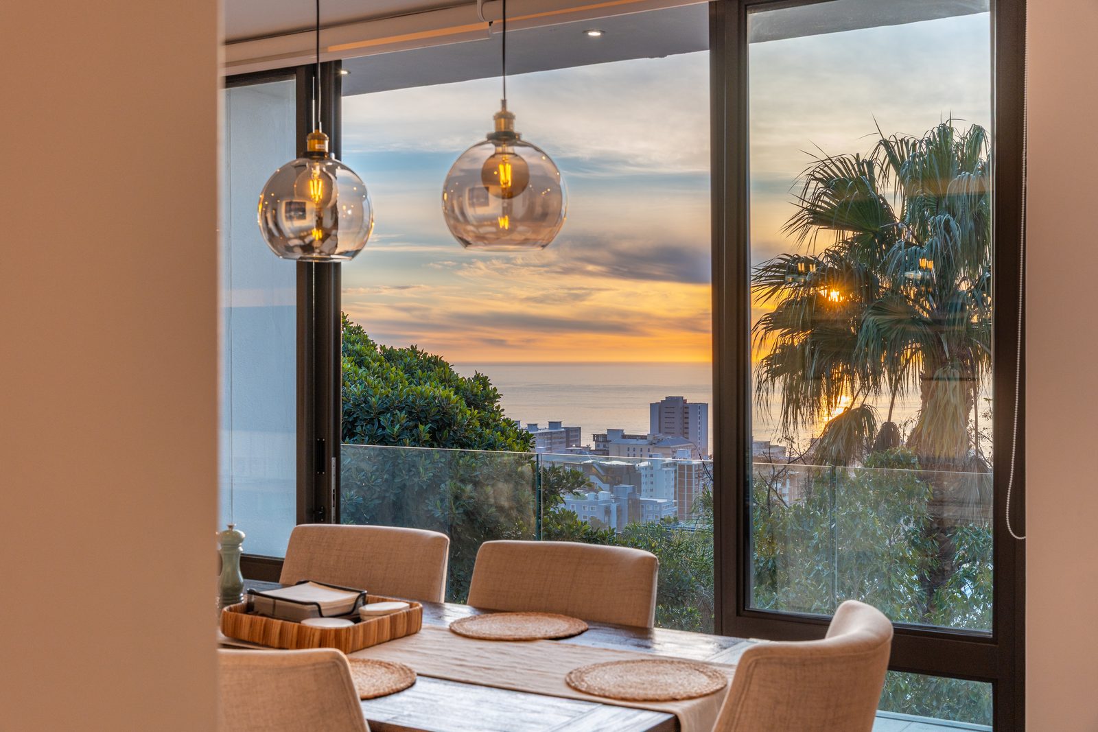 Dining table beneath glass pendant lights with a sunset sea view behind